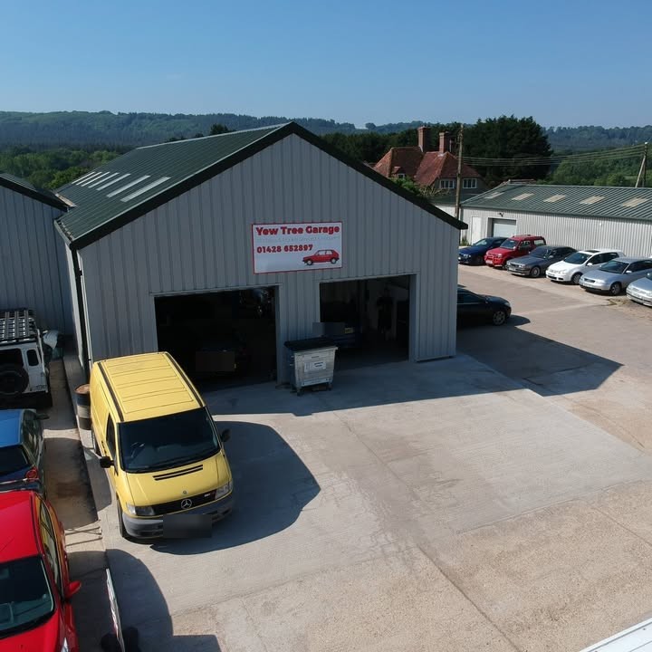 Yew Tree Garage from above: the grey clad unit with its red and white sign, both roller doors open, a yellow van in the yard and the wooded hills behind