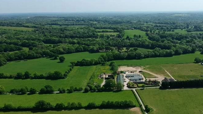 An aerial view of green fields and woodland with a cluster of buildings in the middle distance