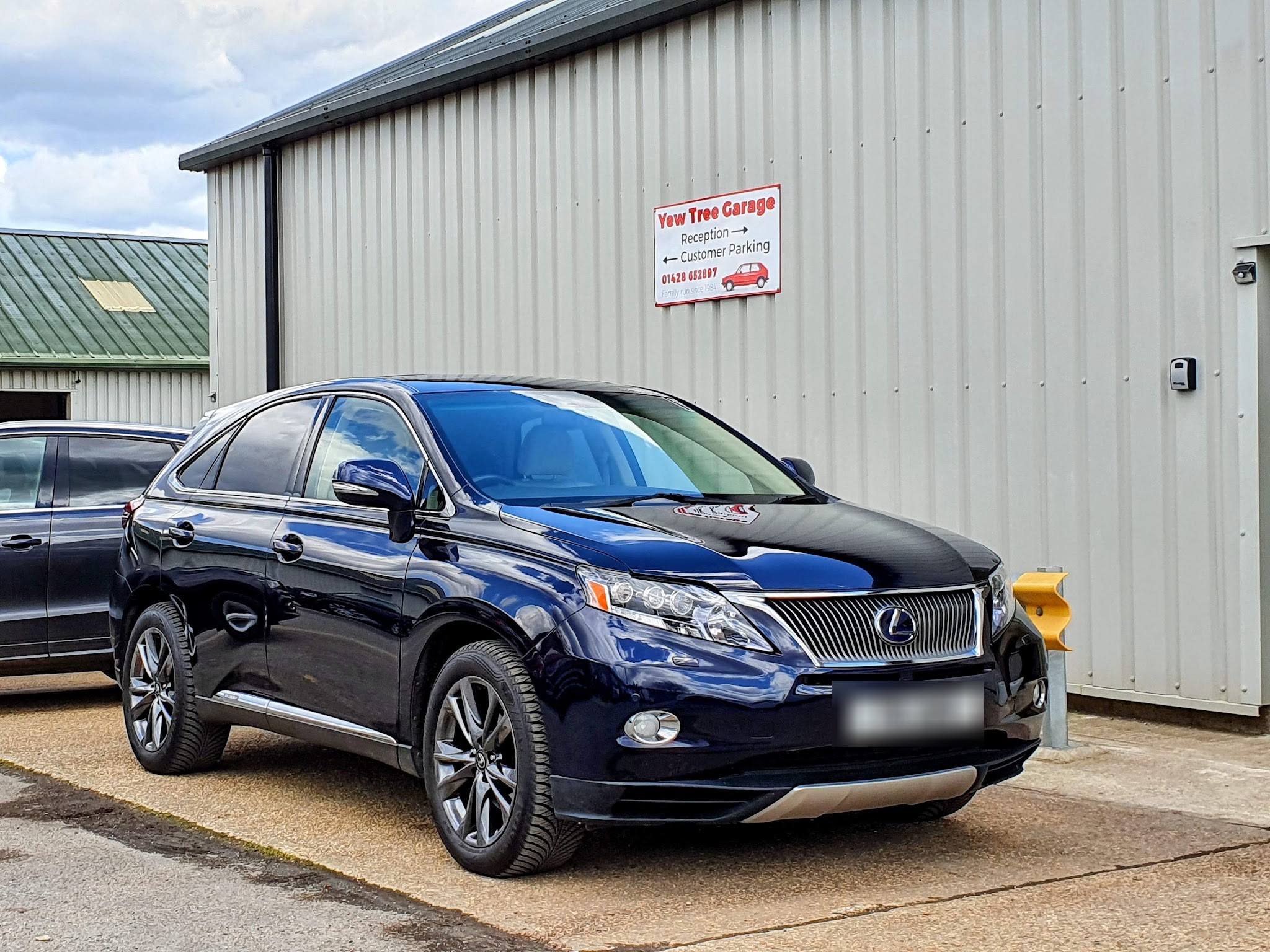 A dark blue Lexus parked outside the garage beside the Yew Tree Garage sign pointing to reception and customer parking
