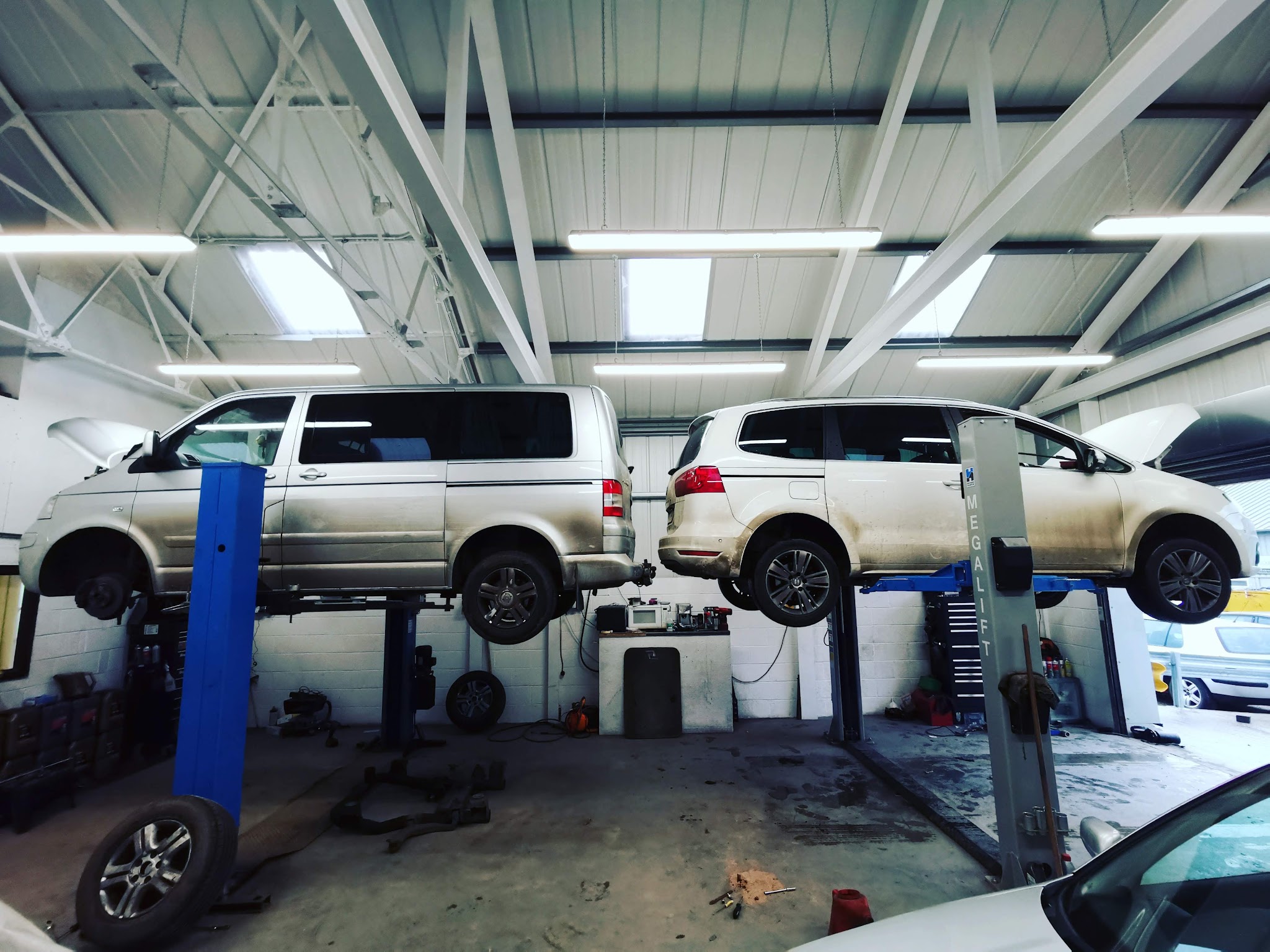 Inside the workshop: a silver VW van and a silver people carrier raised on blue two-post lifts under strip lights