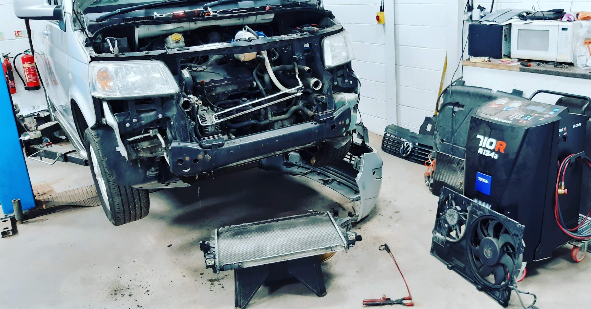 A van in the workshop with its bumper, radiator and fan removed, an air-con service machine beside it